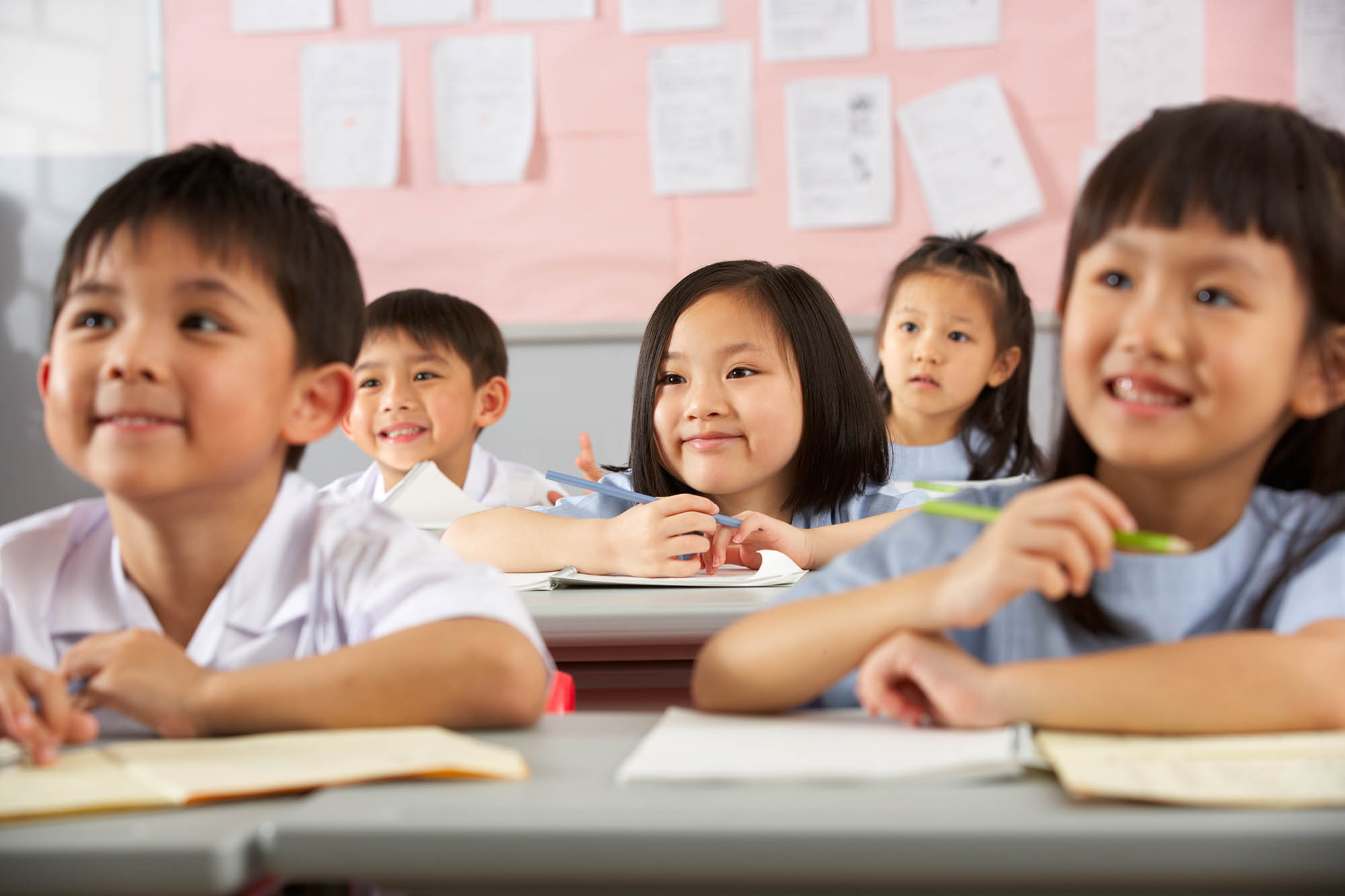 Group Of Students Working At Desks In Chinese School Classroom Looking At Teacher